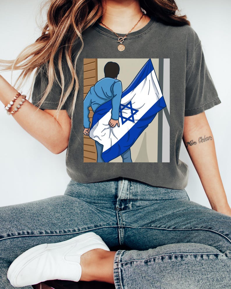 May include: Dark grey t-shirt featuring a graphic of a person holding the Israeli flag. The flag is white with a blue Star of David and blue stripes. The person is wearing a blue jacket and jeans. The shirt is a casual style.