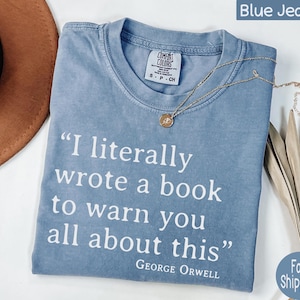 May include: A light blue t-shirt with the quote "I literally wrote a book to warn you all about this" by George Orwell. A gold necklace with a pendant rests on the shirt. The shirt is folded.