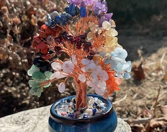 May include: A decorative tree sculpture with a copper wire trunk and branches, adorned with colorful gemstones. The tree sits in a small, blue ceramic pot filled with stones. The gemstones include amethyst, quartz, and other colorful stones.