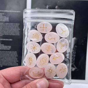 May include: A clear plastic bag filled with 14 pink oval-shaped stones with gold symbols. The stones are in a clear plastic bag with a zipper closure.