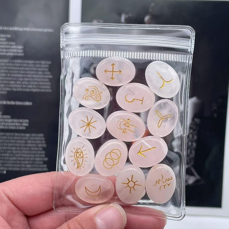 May include: A clear plastic bag filled with 14 pink oval-shaped stones with gold symbols. The stones are in a clear plastic bag with a zipper closure.