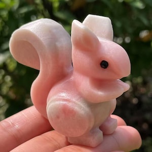 Puede incluir: Una figura de ardilla tallada en rosa y blanco. La ardilla tiene una cola esponjosa, ojos negros pequeños y está en posición de pie. La figura está hecha de una piedra lisa y pulida.