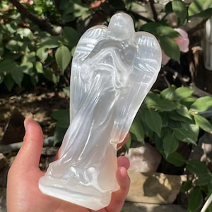 Puede incluir: Una estatua de ángel de selenita blanca con las alas extendidas. El ángel está tallado en una pose de pie con las manos juntas frente al pecho.