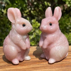 May include: Two pink rabbit figurines with black eyes. The rabbits are made of a sparkly, translucent material and are sitting on a wooden surface.
