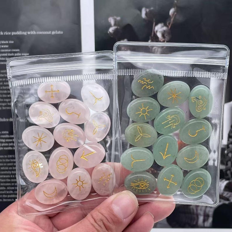 May include: Two clear plastic bags filled with oval-shaped stones. The stones are either pink or green and have gold symbols on them.