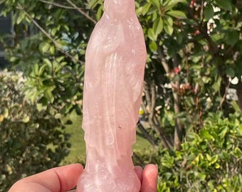 May include: A carved rose quartz statue of a robed figure with hands clasped in prayer. The statue is a soft pink color and stands on a small base. The background is blurred with green foliage.