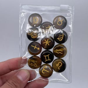 May include: A clear plastic bag containing twelve tiger's eye gemstone zodiac signs. Each stone is round and features a gold zodiac symbol.