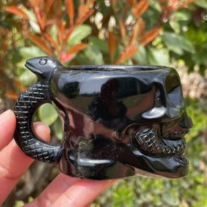 Könnte beinhalten: Schwarzer Obsidian-Schädelbecher mit einem Schlangenhenkel. Der Becher ist poliert und hat ein detailliertes Schädel-Design.