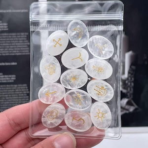 May include: A clear plastic bag filled with white oval-shaped stones. Each stone has a different gold symbol or design on it.
