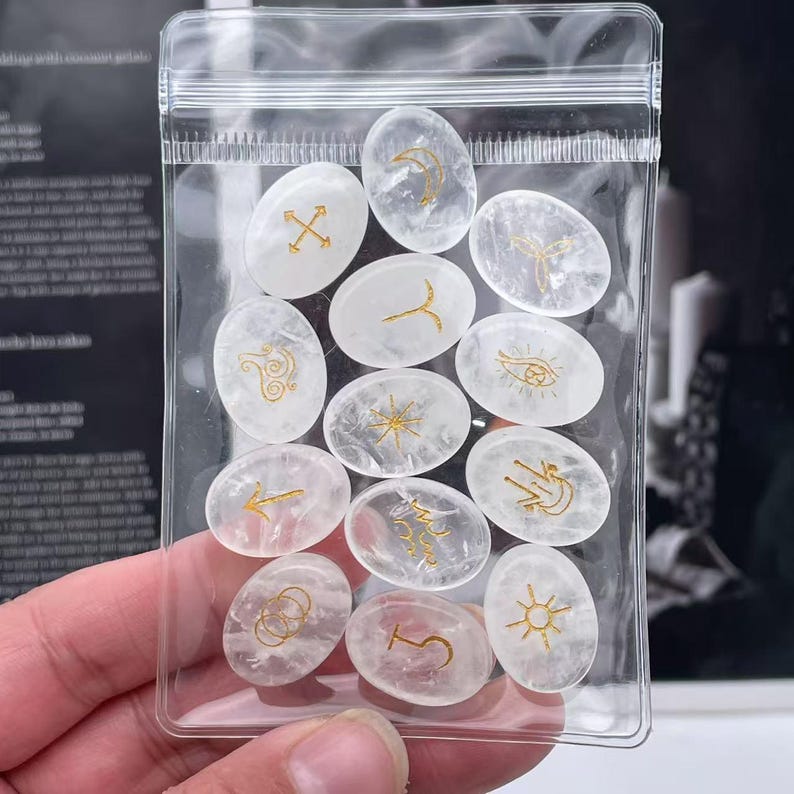 May include: A clear plastic bag filled with white oval-shaped stones. Each stone has a different gold symbol or design on it.