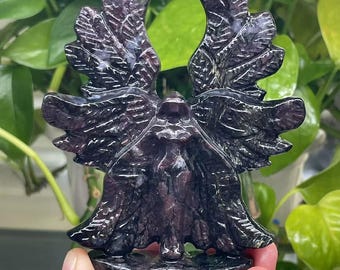 May include: A dark purple carved stone sculpture of a winged creature, possibly an angel or a mythical being. The wings are detailed with feather-like patterns. The sculpture is set against a backdrop of green foliage.