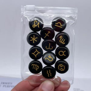 May include: A clear plastic bag filled with twelve black round tokens. Each token has a gold zodiac symbol on it.