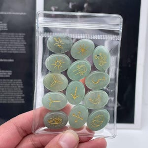 May include: A clear plastic bag filled with 14 green oval-shaped stones with gold symbols. The stones are arranged in two rows.
