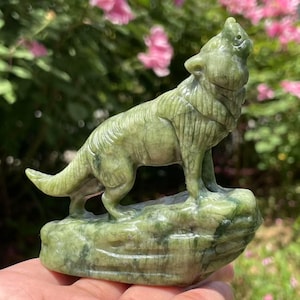 May include: A green stone carving of a howling wolf. The wolf is standing on a rock base, with its head raised and mouth open. The sculpture is detailed, with visible fur and facial features. The stone has a mottled appearance.
