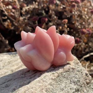 Puede incluir: Un jabón artesanal rosa con forma de flor de loto. El jabón tiene un acabado liso y brillante y está sobre una roca.