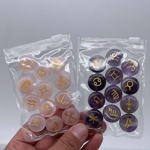 May include: Two clear plastic bags filled with small, round, gemstone zodiac signs. One bag contains pink stones with gold zodiac symbols, and the other bag contains purple stones with gold zodiac symbols.