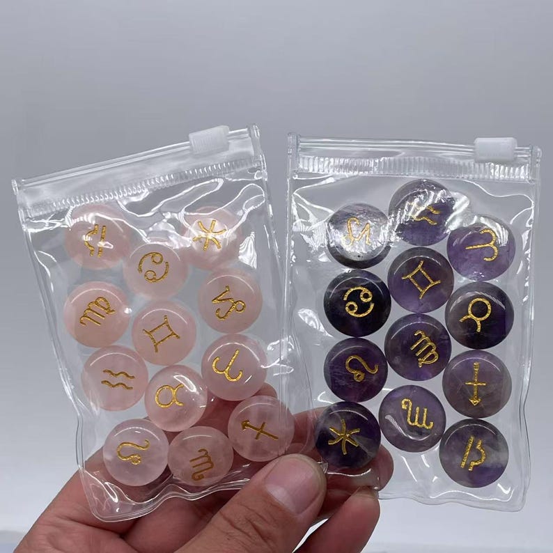 May include: Two clear plastic bags filled with small, round, gemstone zodiac signs. One bag contains pink stones with gold zodiac symbols, and the other bag contains purple stones with gold zodiac symbols.