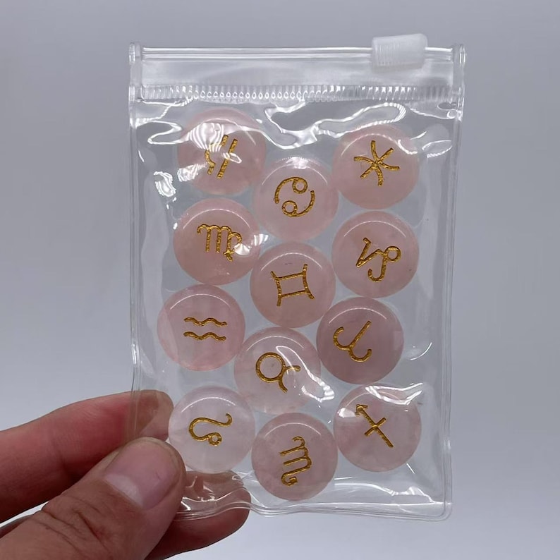 May include: A clear plastic bag containing twelve pink rose quartz stones with gold zodiac symbols. The stones are arranged in a circle.