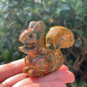 May include: A detailed, hand-carved squirrel figurine. The sculpture features a mix of brown, green, and yellow hues, with a glossy finish. The squirrel is posed with its mouth open, and its tail is curved upwards. The figurine is held in a hand.
