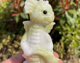 May include: A small, green and white jade dragon figurine with black eyes. The dragon is carved in a whimsical style and has wings and a tail.