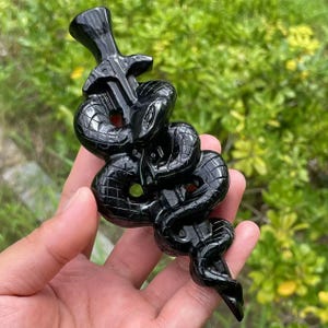 May include: A polished black sculpture depicting a snake coiled around a stylized object. The snake's scales are detailed, and the sculpture has a glossy finish. The background is blurred green foliage.