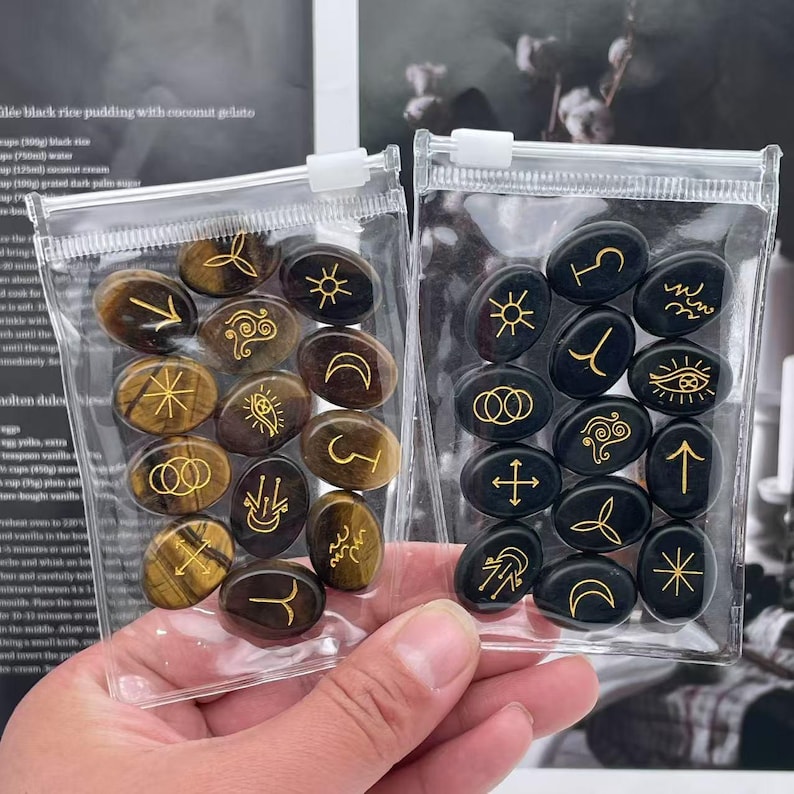 May include: Two clear plastic bags filled with oval-shaped stones. One bag contains tiger's eye stones with gold symbols, and the other bag contains black stones with gold symbols.
