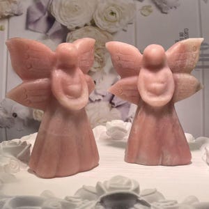 May include: Two pink angel figurines with detailed wings and flowing gowns. The angels are carved from a light pink stone, with a smooth, polished finish. The background features white floral decorations.