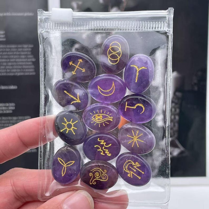 May include: A clear plastic bag filled with 12 small, oval-shaped amethyst stones. Each stone has a different gold symbol etched on the surface.