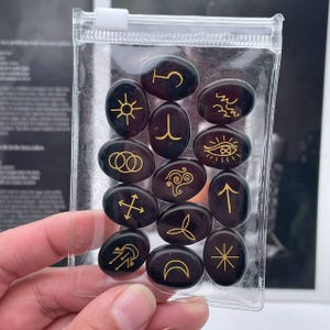 May include: A set of 12 black oval-shaped stones with gold symbols. The stones are in a clear plastic bag.