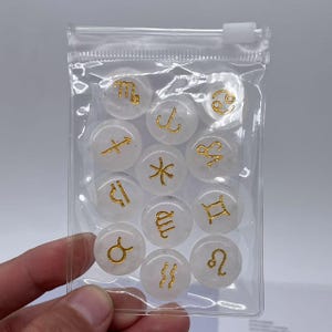 May include: A clear plastic bag containing twelve white round stones with gold zodiac symbols. The stones are arranged in a circle.