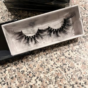May include: A pair of black false eyelashes in a white box with a marble effect. The lashes are long and full, with a natural curl. The box is open, showing the lashes against a white interior.