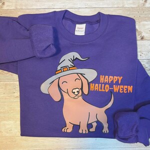 Könnte beinhalten: Lila Sweatshirt mit einem Cartoon-Dackel, der einen Hexenhut trägt. Der Hund ist hellbraun mit einem grauen und orangefarbenen Hut. Die Worte "HAPPY HALLO-WEEN" sind in Orange aufgedruckt. Das Sweatshirt ist aus weichem Material.