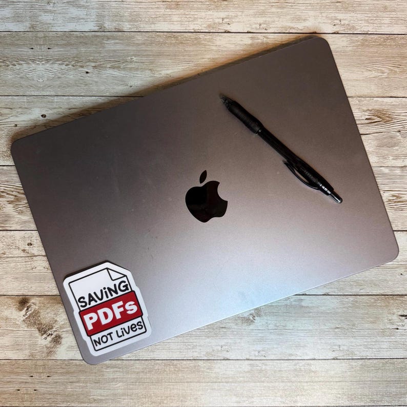 Puede incluir: Un port&aacute;til plateado con un bol&iacute;grafo negro y una pegatina que dice "SAVING PDFs NOT Lives". El port&aacute;til tiene un logotipo de Apple negro. El bol&iacute;grafo es negro y est&aacute; inclinado sobre la parte superior del port&aacute;til.