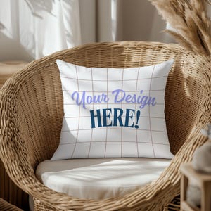 Könnte beinhalten: Ein weißes Kissen mit braunem Gittermuster und dem Text "Your Design HERE!" in blauer und lila Schrift. Das Kissen liegt auf einem Korbstuhl.