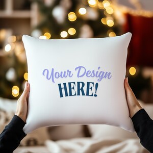 Peut inclure: Un coussin carré blanc avec le texte "Your Design HERE!" en bleu et violet. Le coussin est tenu par une personne. L'arrière-plan est flou avec des lumières de Noël.