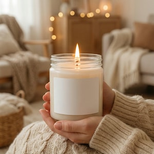 May include: A lit candle in a clear glass jar with a blank white label. The candle flame is bright against the soft, creamy wax. The jar is held in hands wearing a beige sweater, with a cozy, warm background.