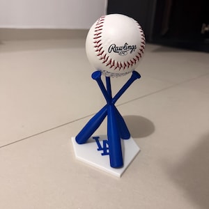 May include: A white baseball with red stitching and the Rawlings logo sits on a blue bat-shaped stand. The stand is on a white base with the Los Angeles Dodgers logo. The baseball is a classic white colour.
