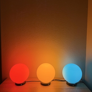 Puede incluir: Tres lámparas LED esféricas que cambian de color se colocan sobre una superficie de madera. Las lámparas se iluminan en rojo, naranja y azul, cada una con una base negra. El fondo es una pared texturizada de tonos neutros.