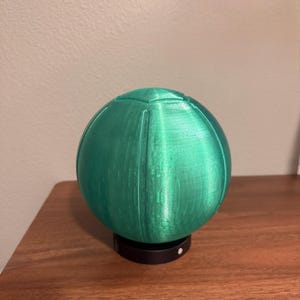 May include: A 3D-printed, spherical object in a vibrant green hue, resting on a small, black circular base. The sphere has visible vertical lines, suggesting a segmented design. The object is displayed on a wooden surface.