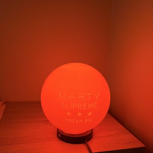 May include: An orange sphere lamp with the text "MARTY SUPREME" and "DREAM BIG" printed on the front. The lamp sits on a black base and is plugged into an electrical outlet. The lamp is on a wooden nightstand.