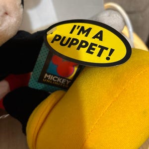 Pu&ograve; includere: Un burattino di peluche di Topolino con una scarpa giallo brillante e un corpo nero. Un'etichetta ovale gialla attaccata al burattino mostra il testo nero I'M A PUPPET! sopra un logo Mickey Unlimited con una sagoma rossa delle orecchie di Topolino.