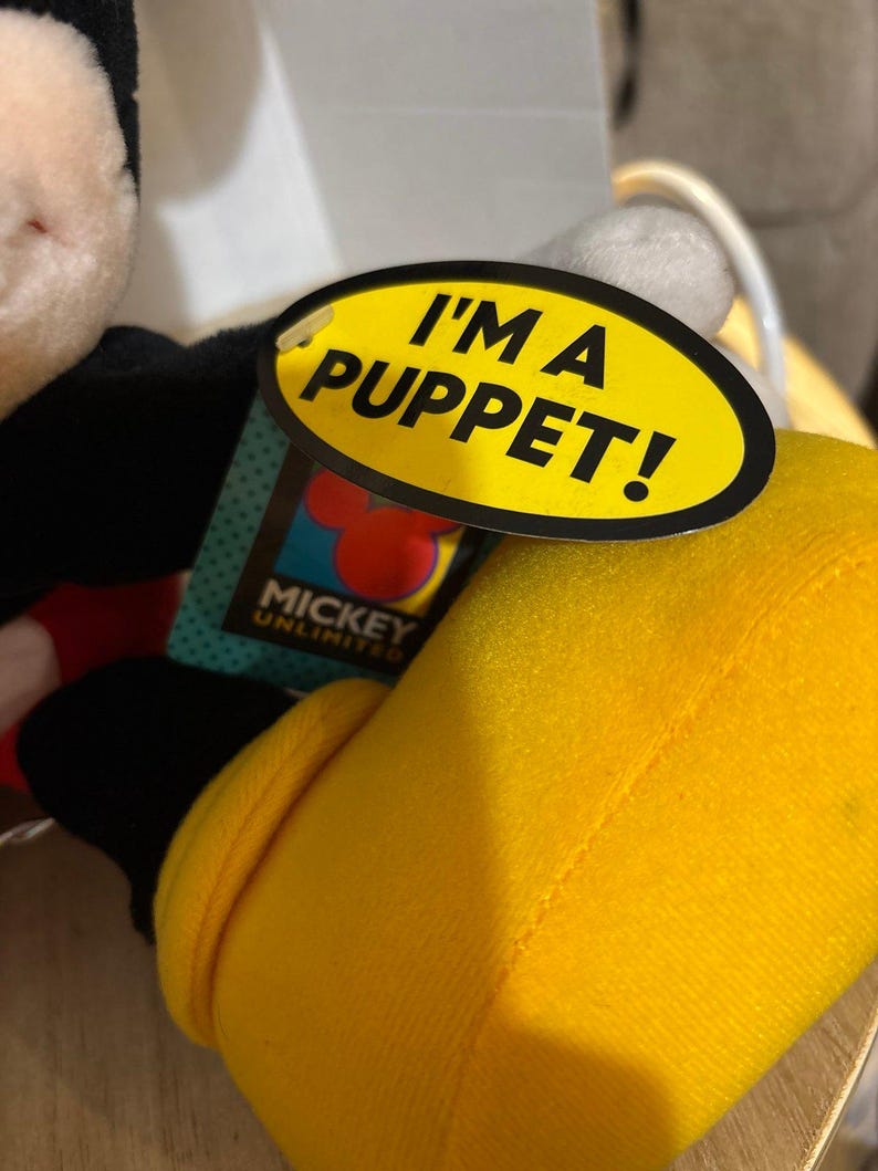 Pu&ograve; includere: Un burattino di peluche di Topolino con una scarpa giallo brillante e un corpo nero. Un'etichetta ovale gialla attaccata al burattino mostra il testo nero I'M A PUPPET! sopra un logo Mickey Unlimited con una sagoma rossa delle orecchie di Topolino.