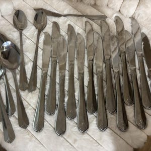 May include: A collection of stainless steel flatware including several spoons and knives. Each piece features a polished silver finish and a decorative fan-shaped detail at the base of the handle. The set consists of multiple matching utensils.
