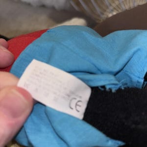 Puede incluir: Un peluche con una camisa de tela azul brillante y una secci&oacute;n de tela roja. Una etiqueta de cuidado rectangular blanca est&aacute; unida a la tela azul, mostrando texto impreso y una marca CE. Se ve una extremidad de tela negra bajo la camisa azul.