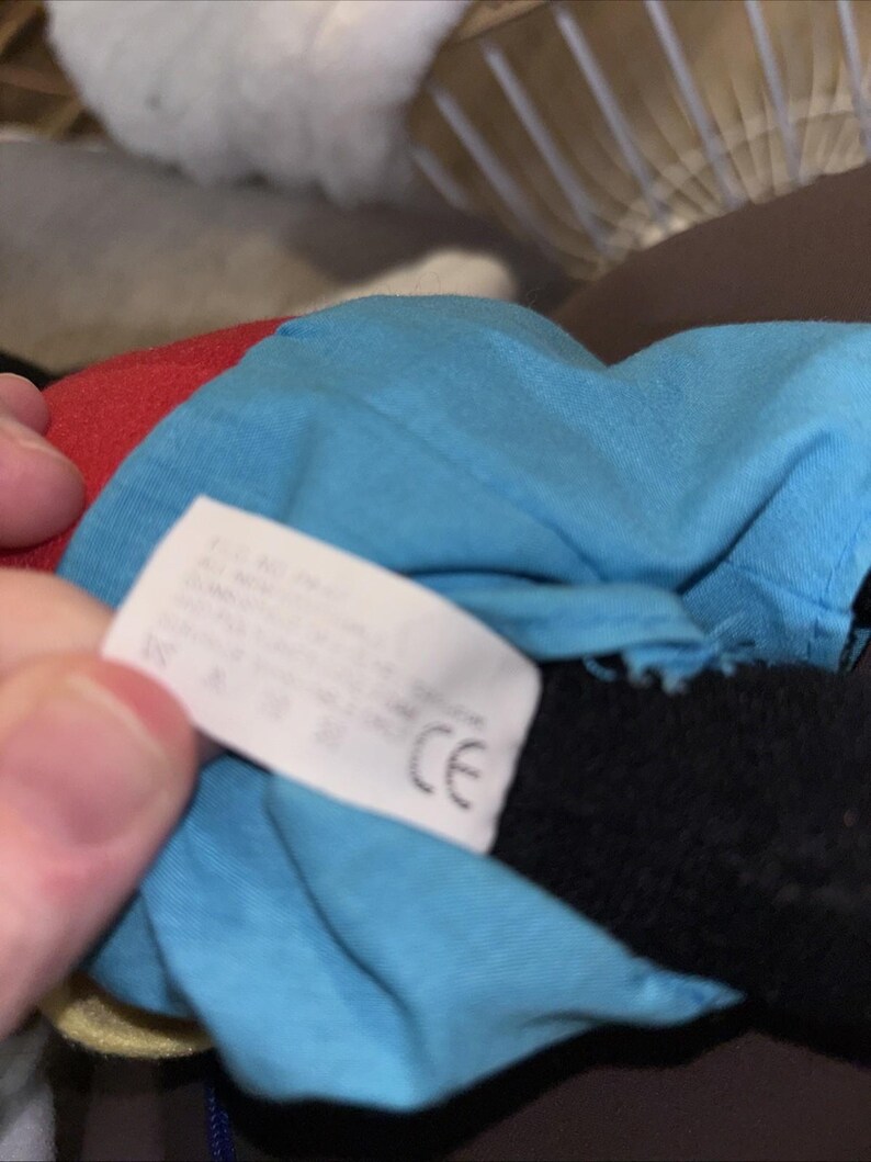 Puede incluir: Un peluche con una camisa de tela azul brillante y una secci&oacute;n de tela roja. Una etiqueta de cuidado rectangular blanca est&aacute; unida a la tela azul, mostrando texto impreso y una marca CE. Se ve una extremidad de tela negra bajo la camisa azul.