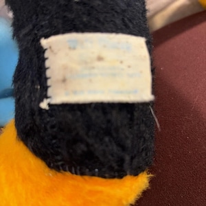 May include: Close-up of a plush toy's leg. The toy has black fur on the leg and a bright orange foot. A small, rectangular tag is attached to the leg, with faded text.