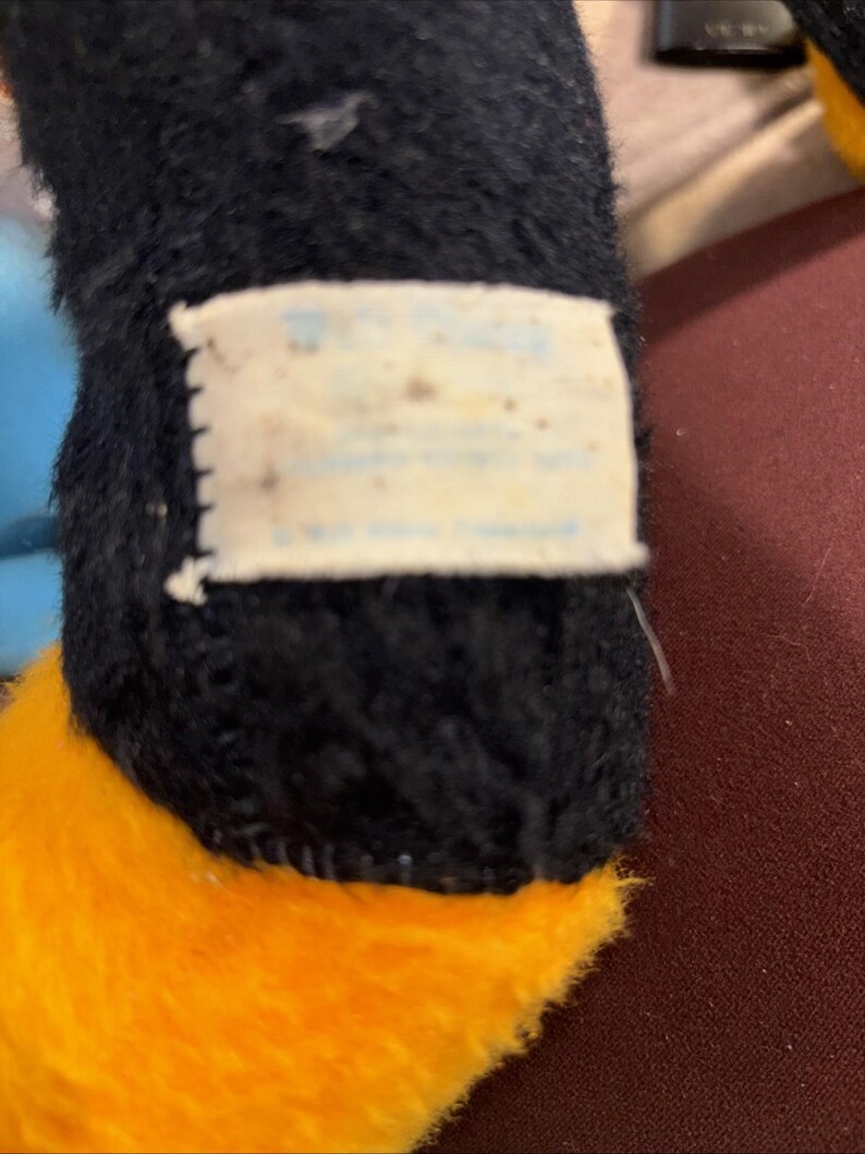 May include: Close-up of a plush toy's leg. The toy has black fur on the leg and a bright orange foot. A small, rectangular tag is attached to the leg, with faded text.