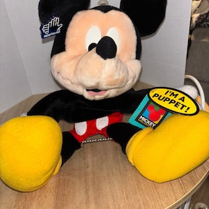 Pu&ograve; includere: Un burattino di peluche di Topolino con pelliccia nera, viso color pesca, pantaloncini rossi con due bottoni bianchi e grandi scarpe gialle. Il pupazzo include un'etichetta del marchio Applause e una gialla con scritto I'M A PUPPET! MICKEY UNLIMITED.
