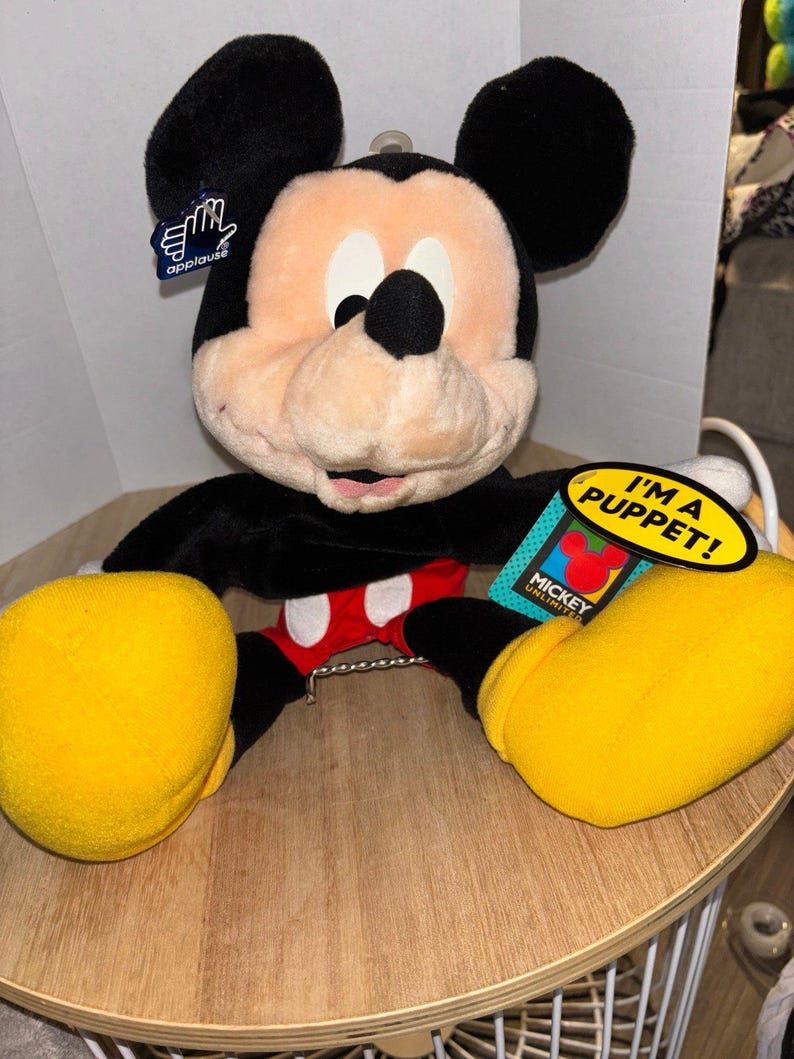 Pu&ograve; includere: Un burattino di peluche di Topolino con pelliccia nera, viso color pesca, pantaloncini rossi con due bottoni bianchi e grandi scarpe gialle. Il pupazzo include un'etichetta del marchio Applause e una gialla con scritto I'M A PUPPET! MICKEY UNLIMITED.