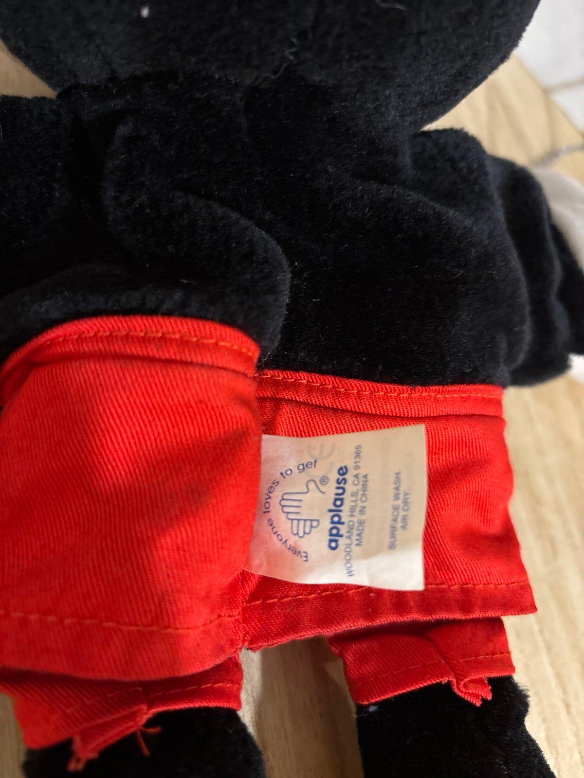 Pu&ograve; includere: Un peluche nero che indossa pantaloncini rosso brillante. Un'etichetta in tessuto bianco &egrave; attaccata alla vita, con il testo "applause", "WOODLAND HILLS, CA 91365", "MADE IN CHINA", "SURFACE WASH", "AIR DRY" e "Everyone loves to get" con un'icona a mano.
